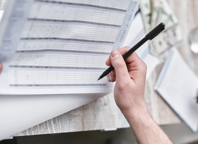A close-up of a person holding a pen reviewing a financial document with cash visible, ideal for business themes. Marketing agencies risk scenarios without the panic buying